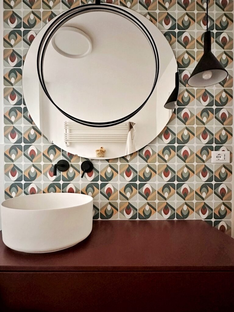 bagno principale di una casa appena ristrutturata su mio progetto che vede un rivestimento con piastrella con fantasia anni 70 geometrica e colorata, lavabo a ciotola, rubinetteria nera e una luce a sospensione nera, specchio tondo con dettaglio circolare nero sovrapposto, tutto segue il progetto di interior design
